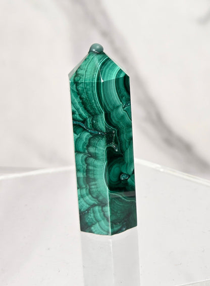 A polished green malachite crystal tower with natural banding patterns, displayed on a clear acrylic hexagonal stand against a white marble background.