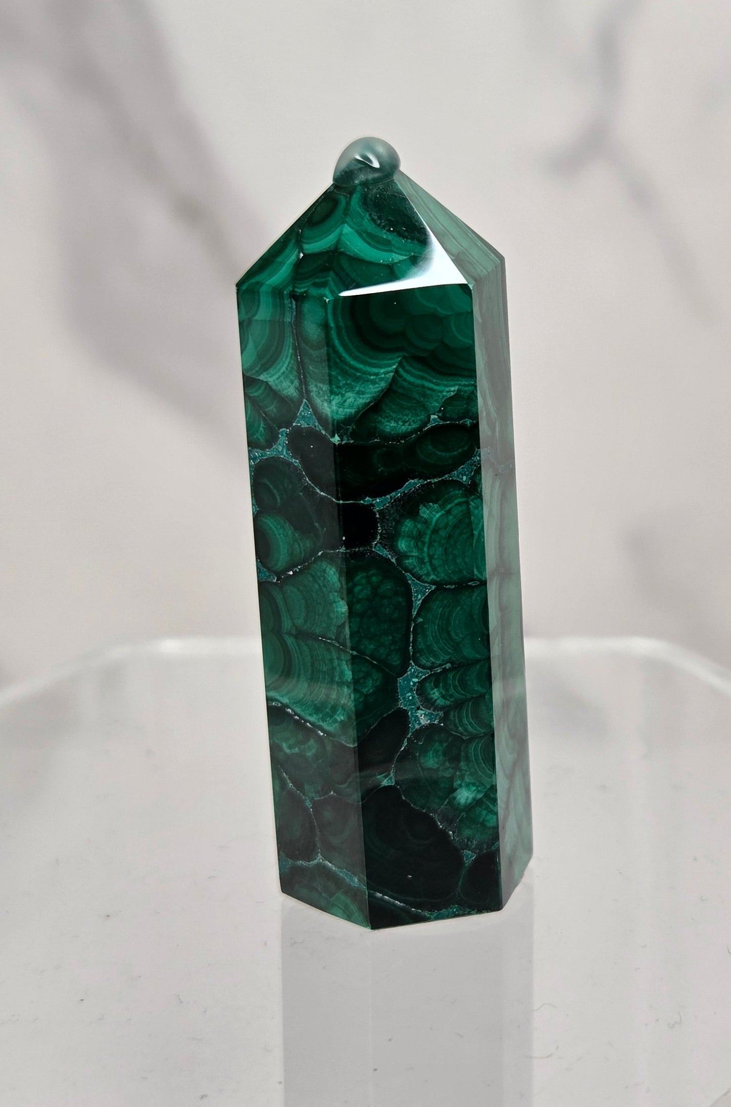 A polished green malachite crystal tower with natural dark veining, displayed on a clear acrylic stand against a light gray marble-patterned background.