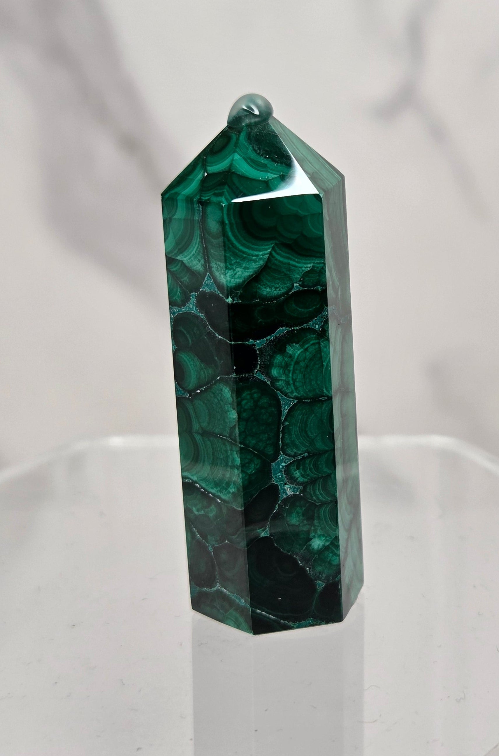 A polished green malachite crystal tower with natural dark veining, displayed on a clear acrylic stand against a light gray marble-patterned background.