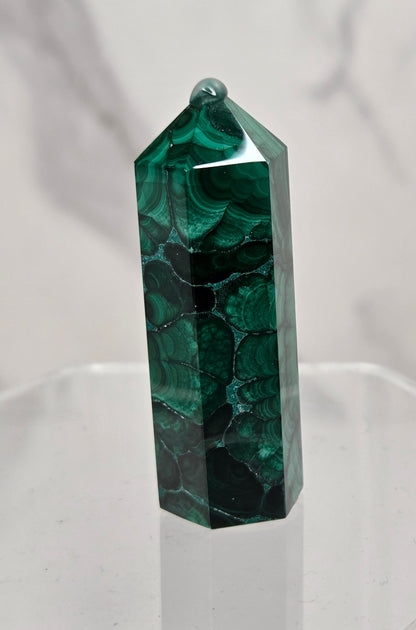 A polished green malachite crystal tower with natural dark veining, displayed on a clear acrylic stand against a light gray marble-patterned background.