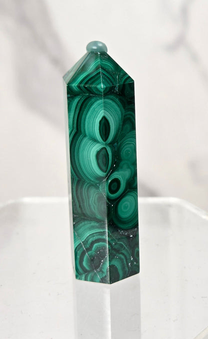 A polished green malachite crystal obelisk with natural banded patterns, displayed on a clear acrylic stand against a light gray marble background.