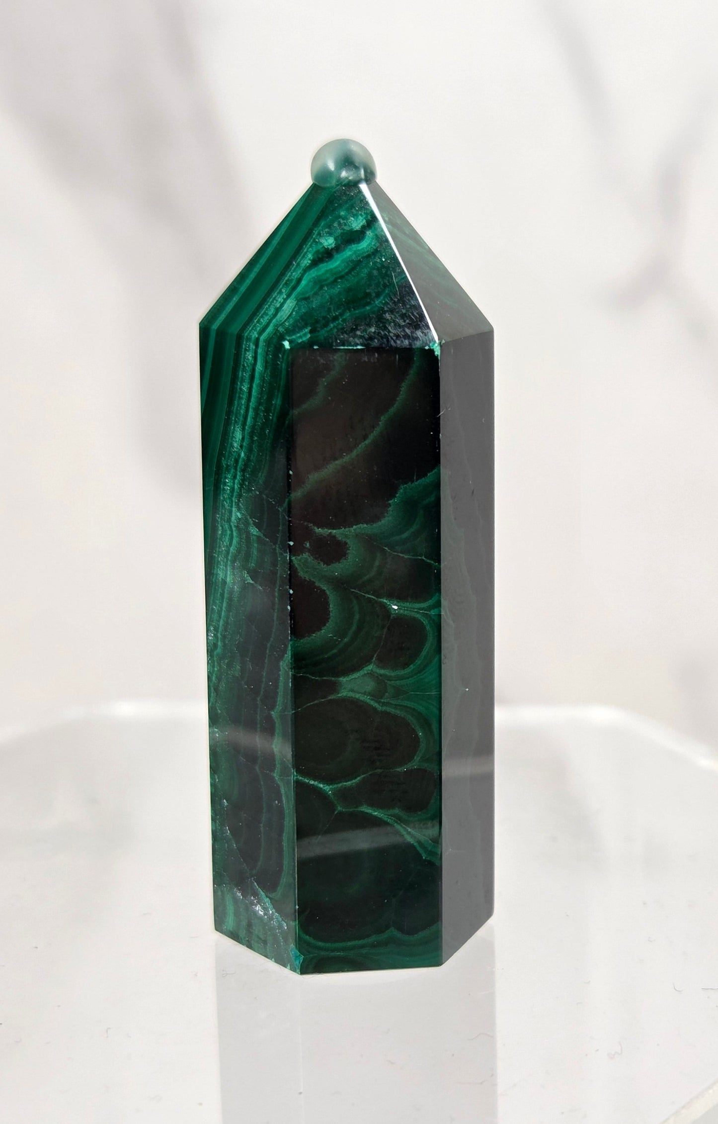 A polished green malachite crystal tower with natural dark green and black marbling, displayed on a clear acrylic stand against a light marble-patterned background.