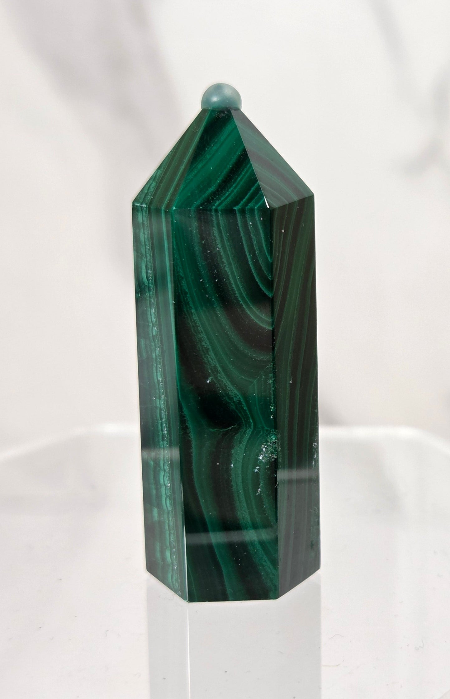 A polished green malachite crystal tower with natural dark and light green banded patterns, displayed on a clear acrylic stand against a white marble background.