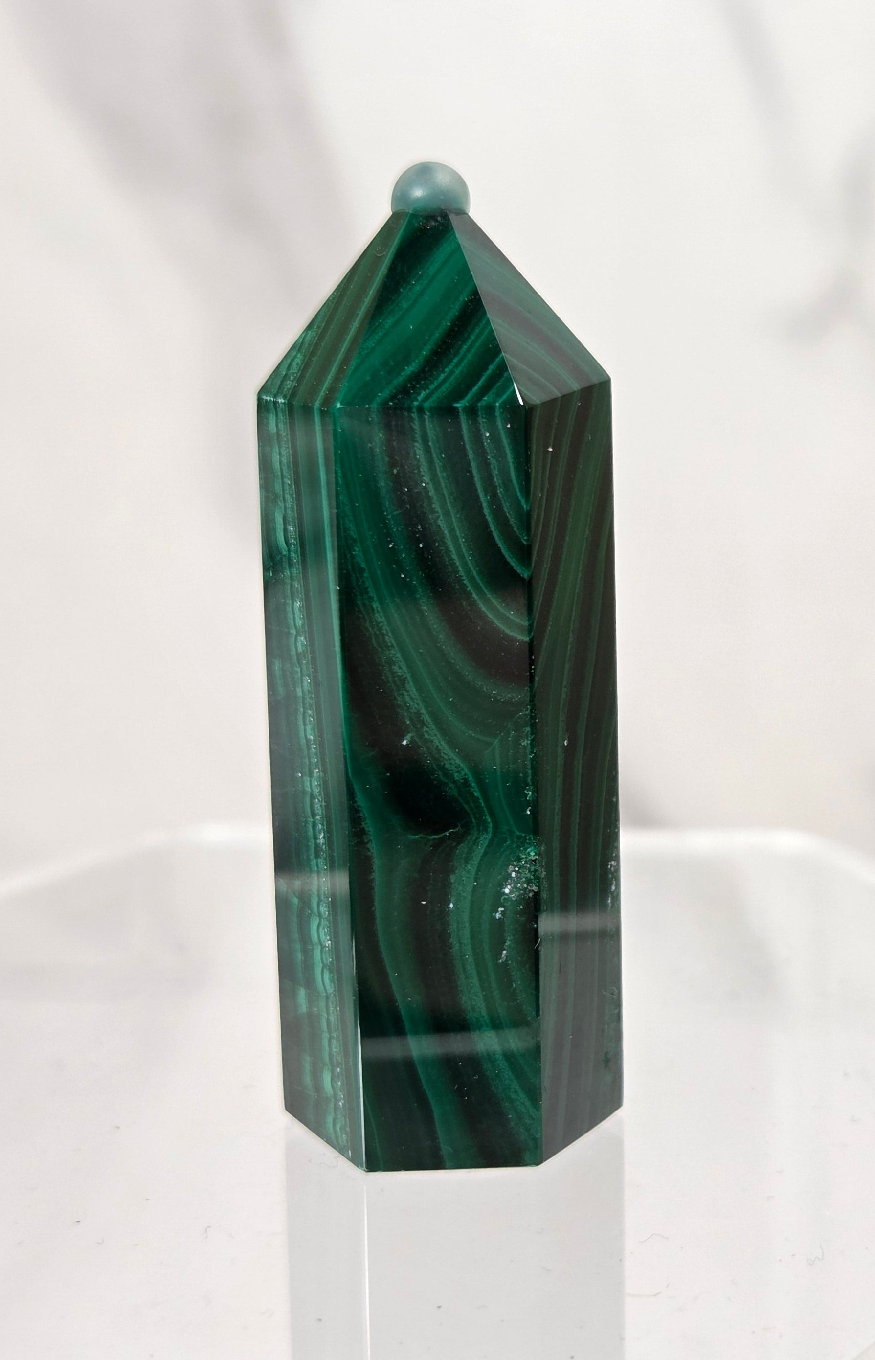 A polished green malachite crystal tower with natural dark and light green banded patterns, displayed on a clear acrylic stand against a white marble background.