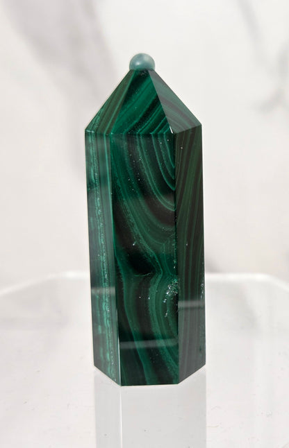 A polished green malachite crystal tower with natural dark and light green banded patterns, displayed on a clear acrylic stand against a white marble background.