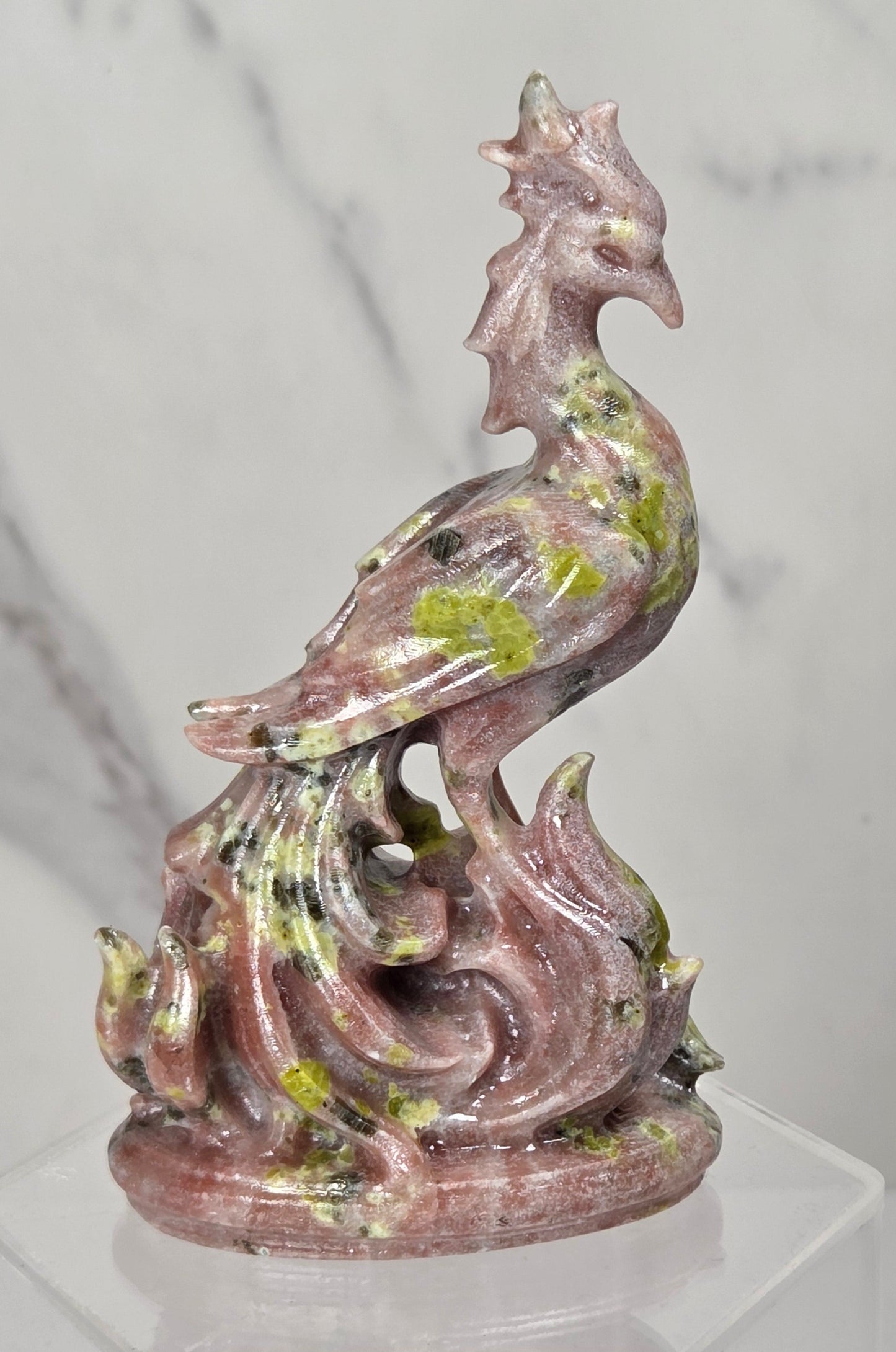 A carved stone figurine of a phoenix rising from flames, made from pinkish-red jasper with green and yellowish inclusions, displayed on a clear acrylic stand against a white marble background.