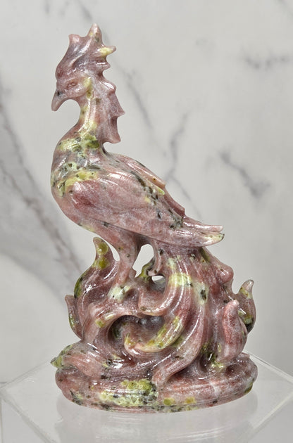 A pink and green speckled stone sculpture of a rooster standing on a flame-like base, displayed on a clear acrylic stand against a white marble-patterned background.