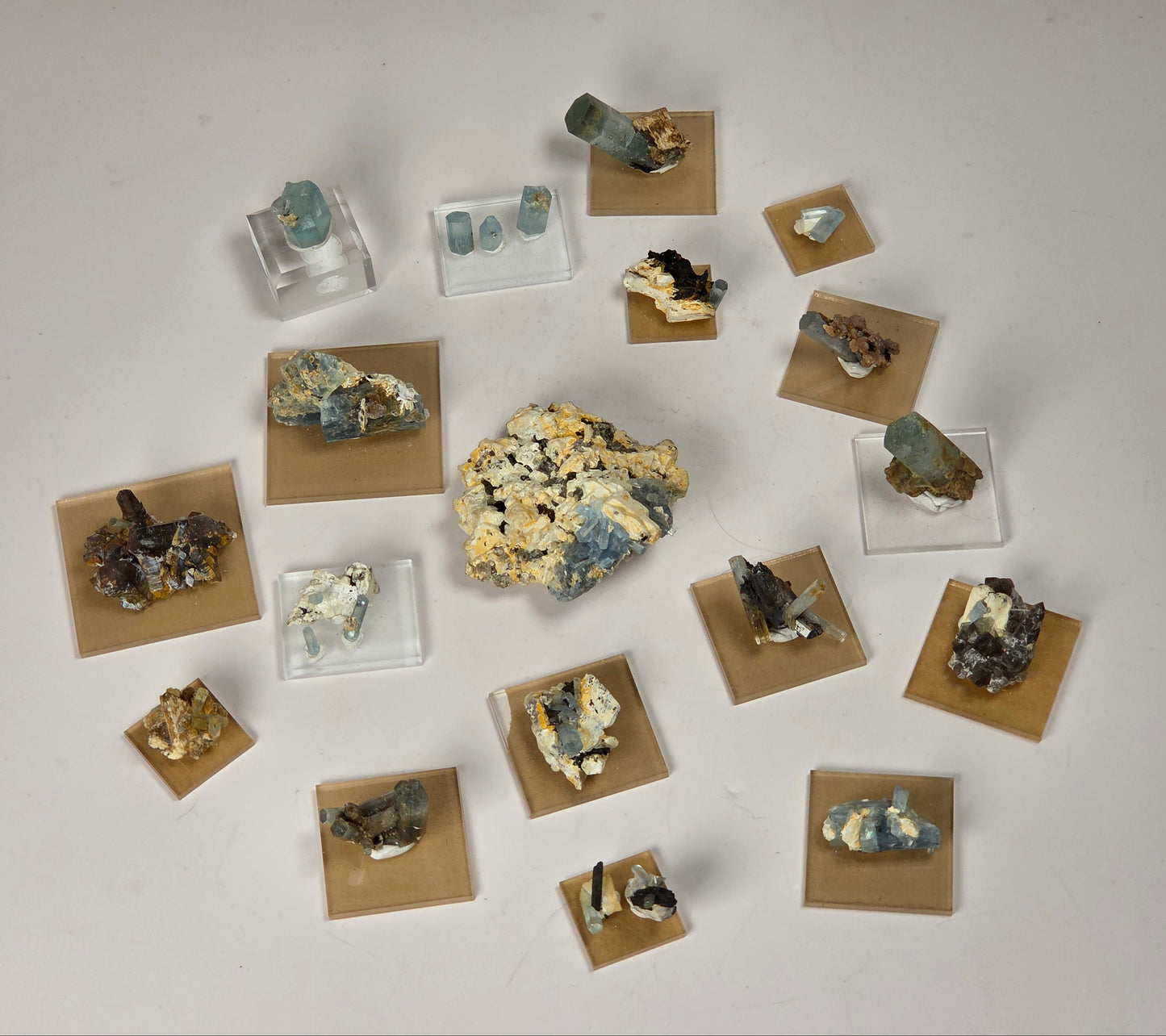 An assortment of mineral and gemstone specimens displayed on small clear acrylic stands and tan square cards against a plain light gray background.