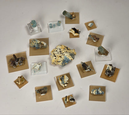 An assortment of mineral and gemstone specimens displayed on small clear acrylic stands and tan square cards against a plain light gray background.