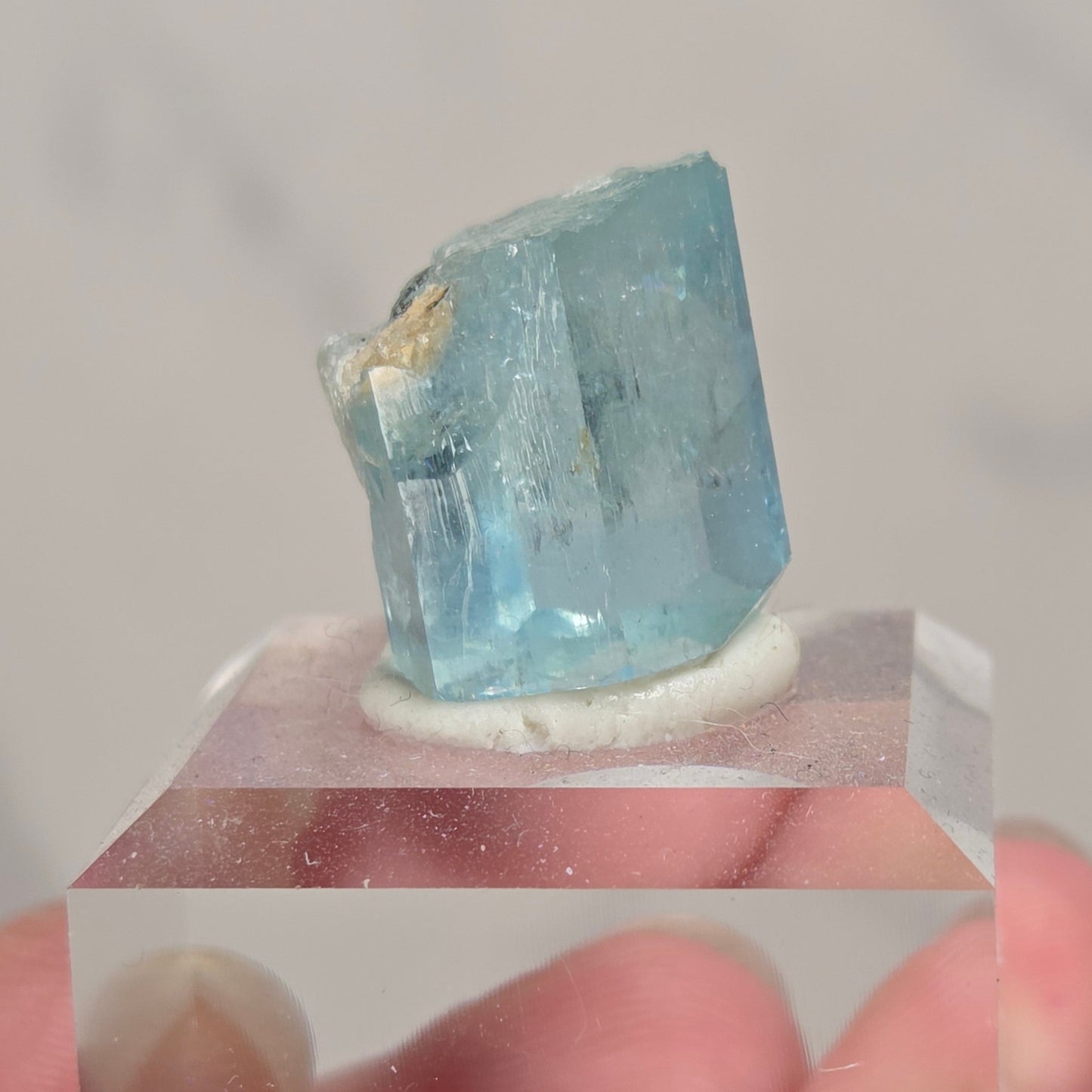 A hand holding a small faceted light blue crystal mounted on a clear acrylic block against a soft light gray background.