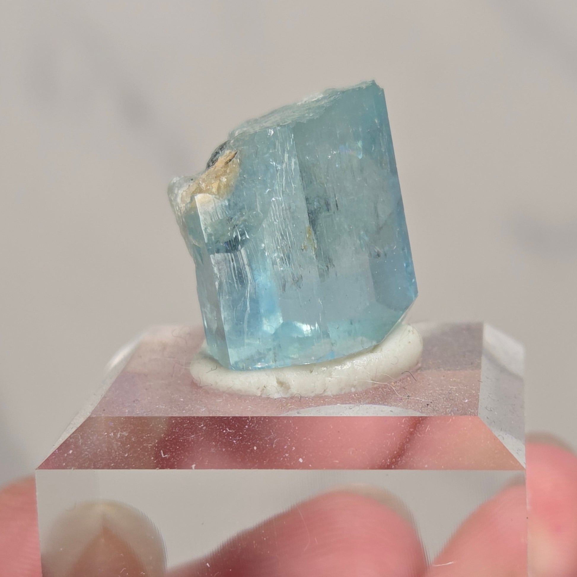 A hand holding a small faceted light blue crystal mounted on a clear acrylic block against a soft light gray background.