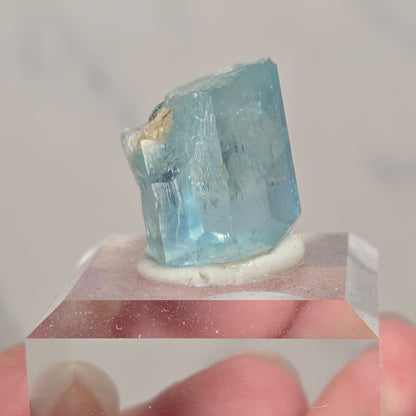 A hand holding a small faceted light blue crystal mounted on a clear acrylic block against a soft light gray background.