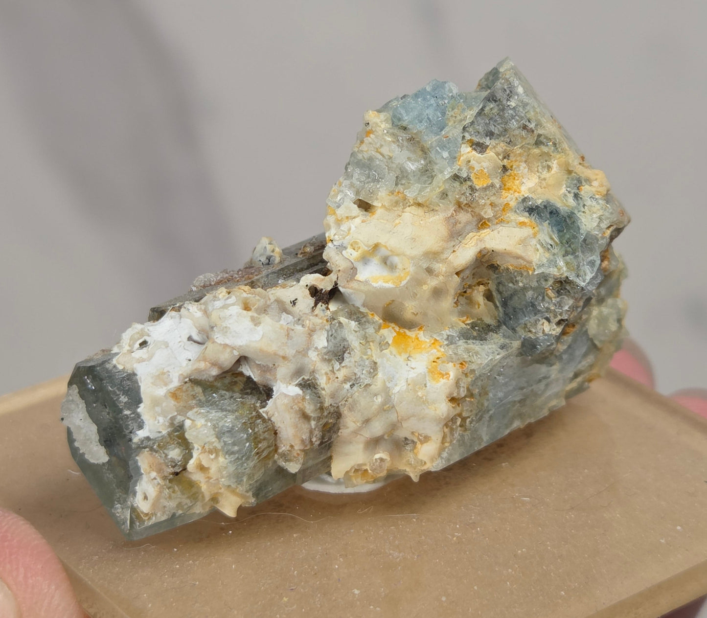 A small mineral specimen with blue-gray, cream, and yellow-orange crystalline texture mounted on a tan display block, held in a person's hand against a soft gray marble-like background.