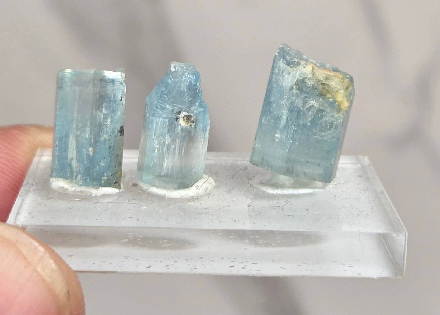 Three small light blue translucent crystal points mounted on a clear acrylic display block, held between fingers against a soft gray marble-patterned background.