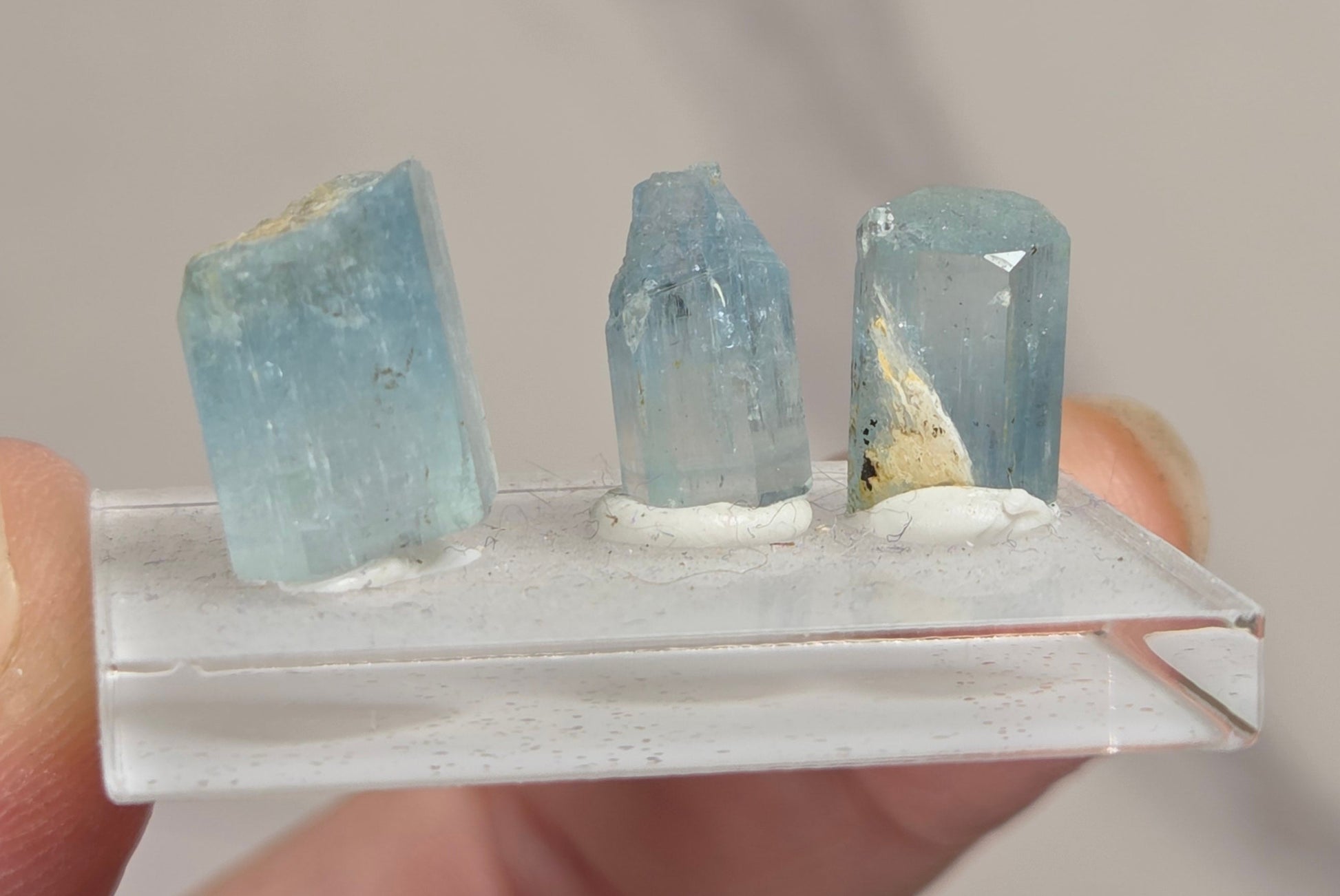 Three small light blue gemstone point specimens mounted on a clear acrylic display block, held between fingers against a soft light gray background.