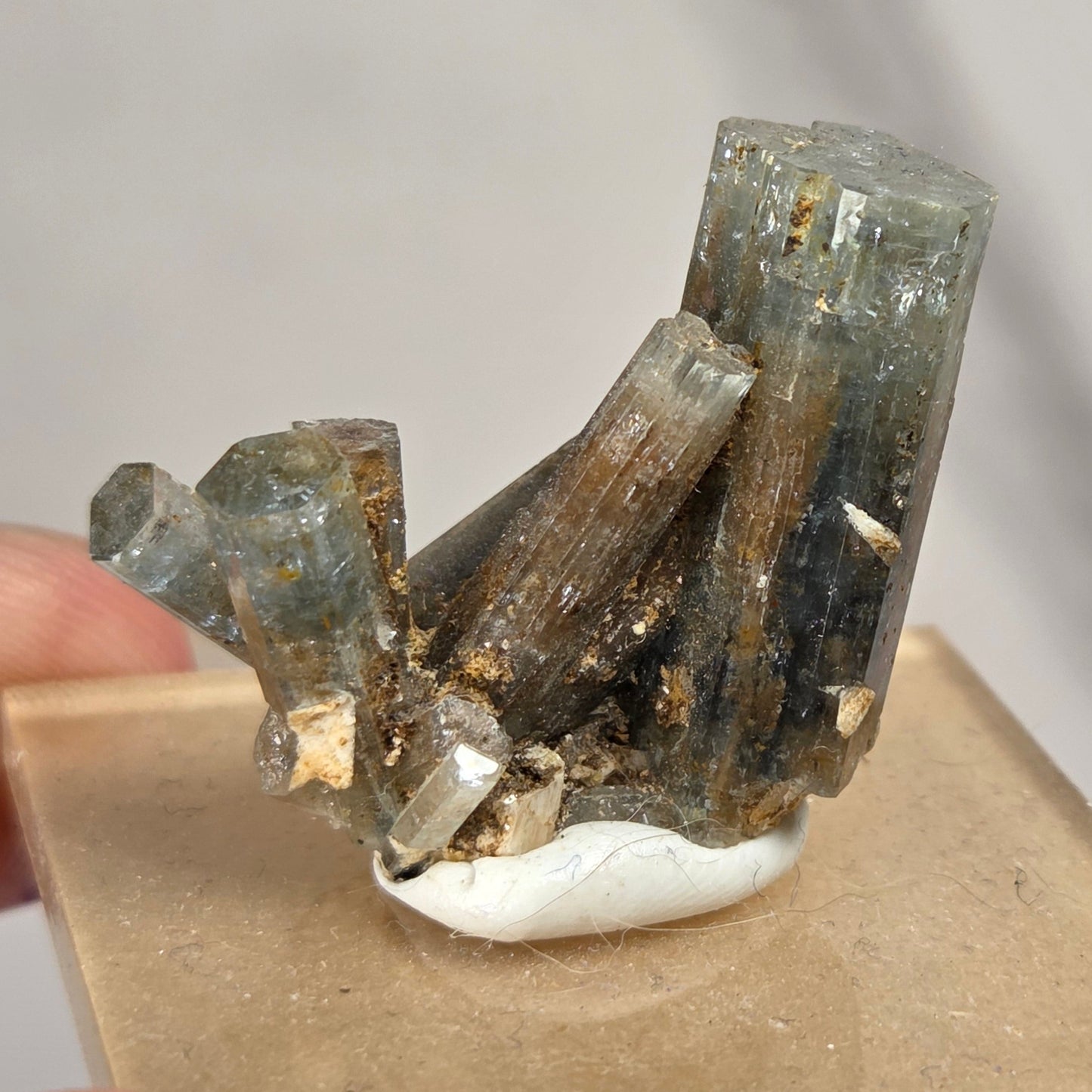 A natural crystal cluster with tall gray and brown translucent mineral points mounted on a small white base and set in a square tan display block, held by fingers against a light gray background.