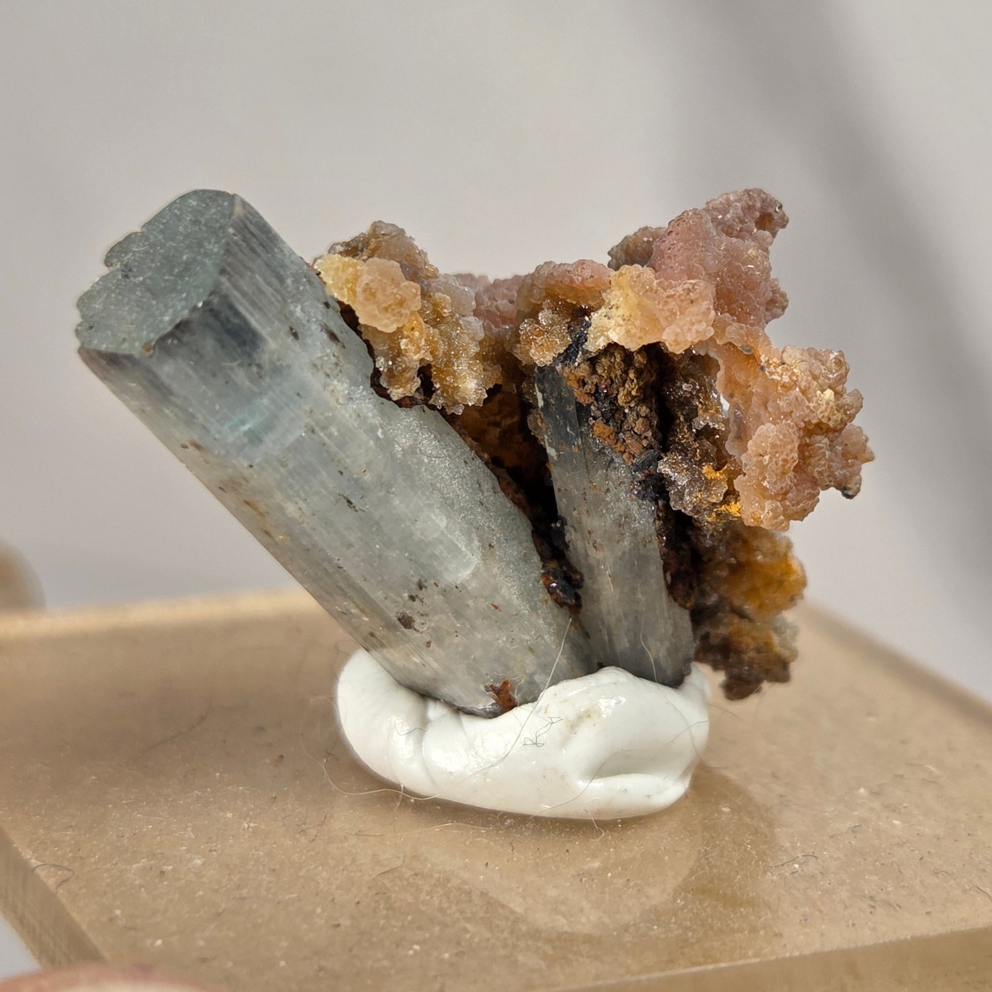 A mineral specimen featuring a gray-blue columnar crystal and orange-brown crystalline growth, mounted on a white support over a tan card, with a person's fingers visible at the bottom left.