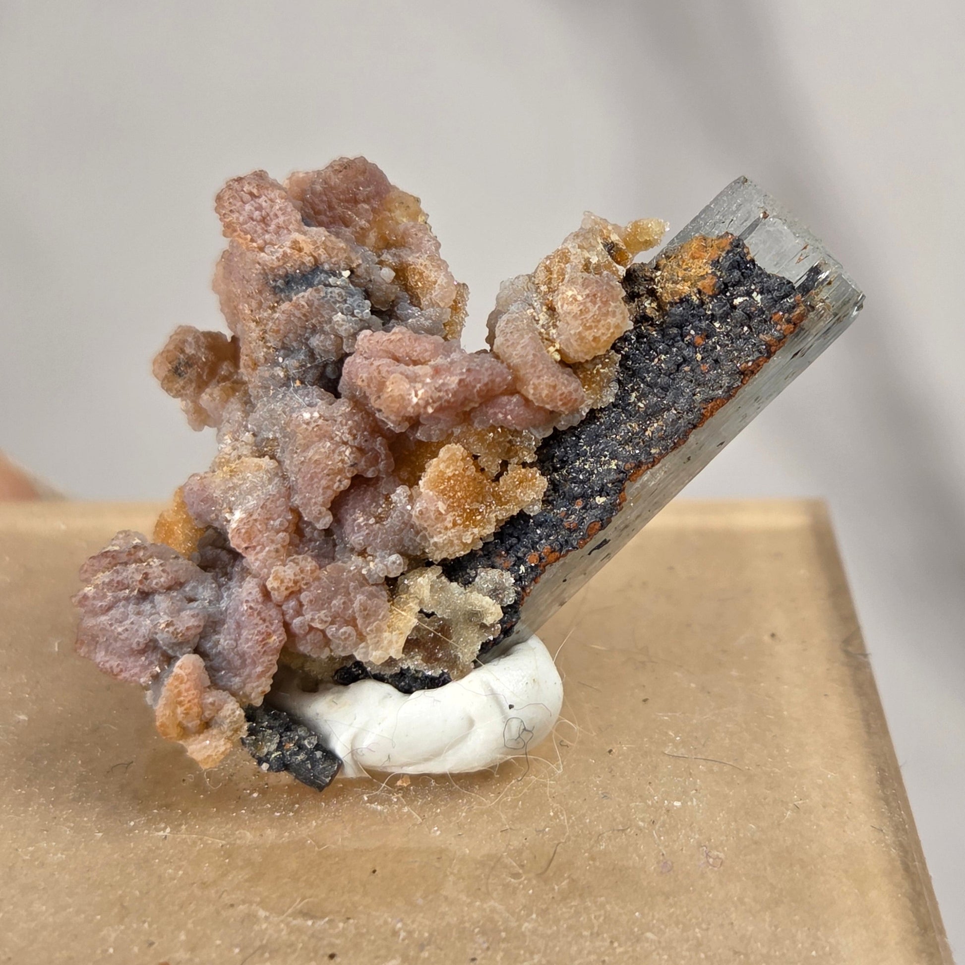 A mineral specimen with pinkish-brown and orange crystalline formations, a dark gray to black matrix, and a white base, mounted on a square tan display block.