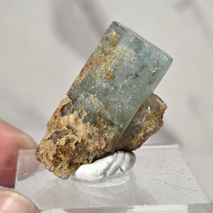 A hand holding a natural mineral specimen featuring a pale blue-green translucent crystal on a rough brown matrix, mounted on a clear acrylic display block.