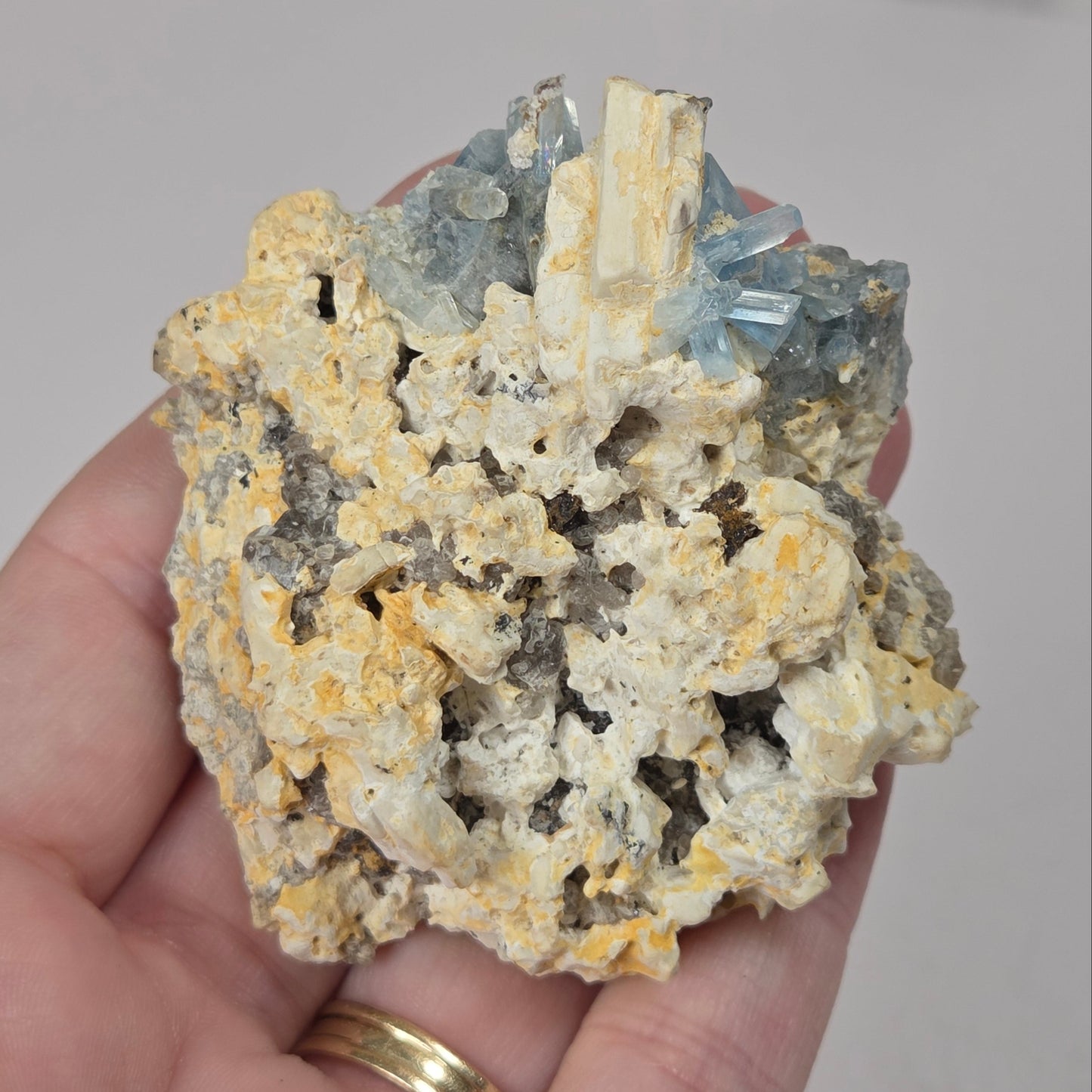 A hand holding a mineral specimen featuring pale yellow calcite crystals, grayish-brown matrix, and light blue quartz points, with a gold ring on the ring finger.
