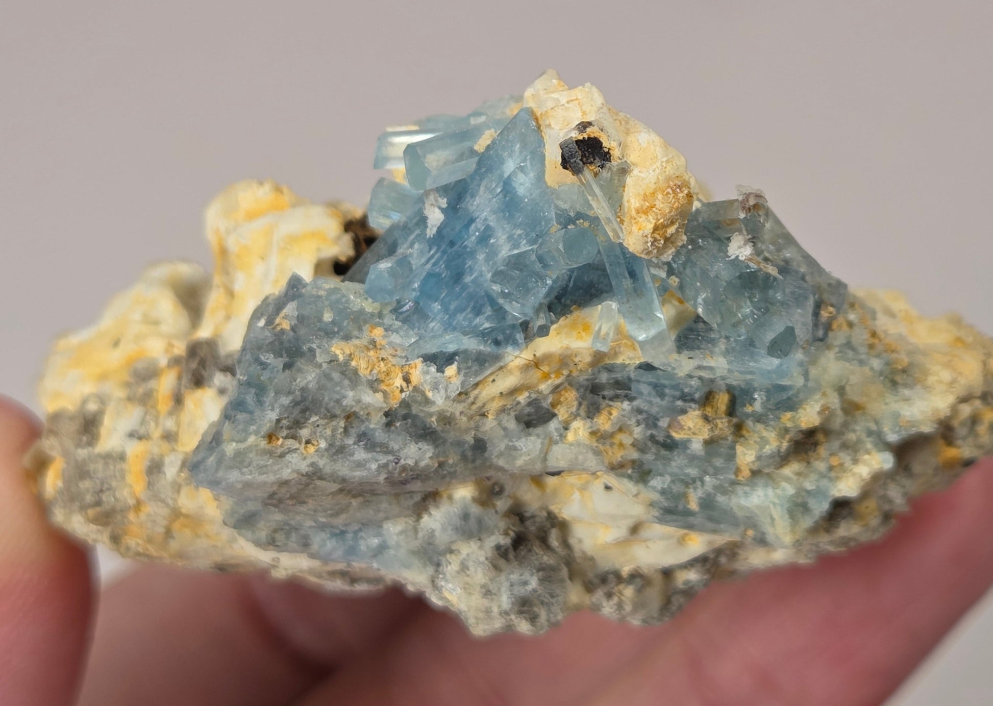 A hand holding a raw mineral specimen featuring light blue fluorite crystals on a yellow and gray matrix, with a blurred neutral background.