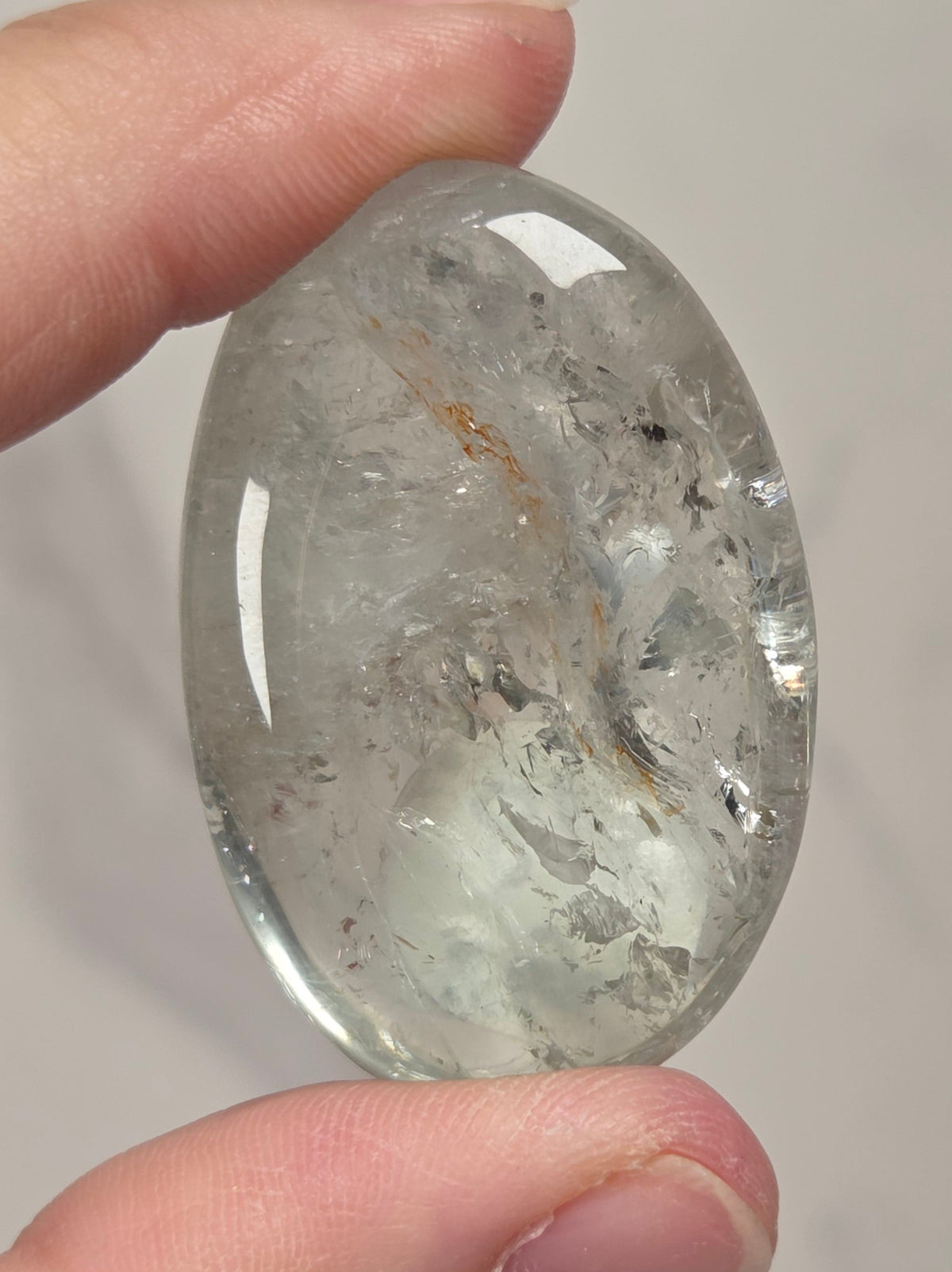 A hand holding a polished oval-shaped clear quartz crystal with internal inclusions against a light gray marble background.