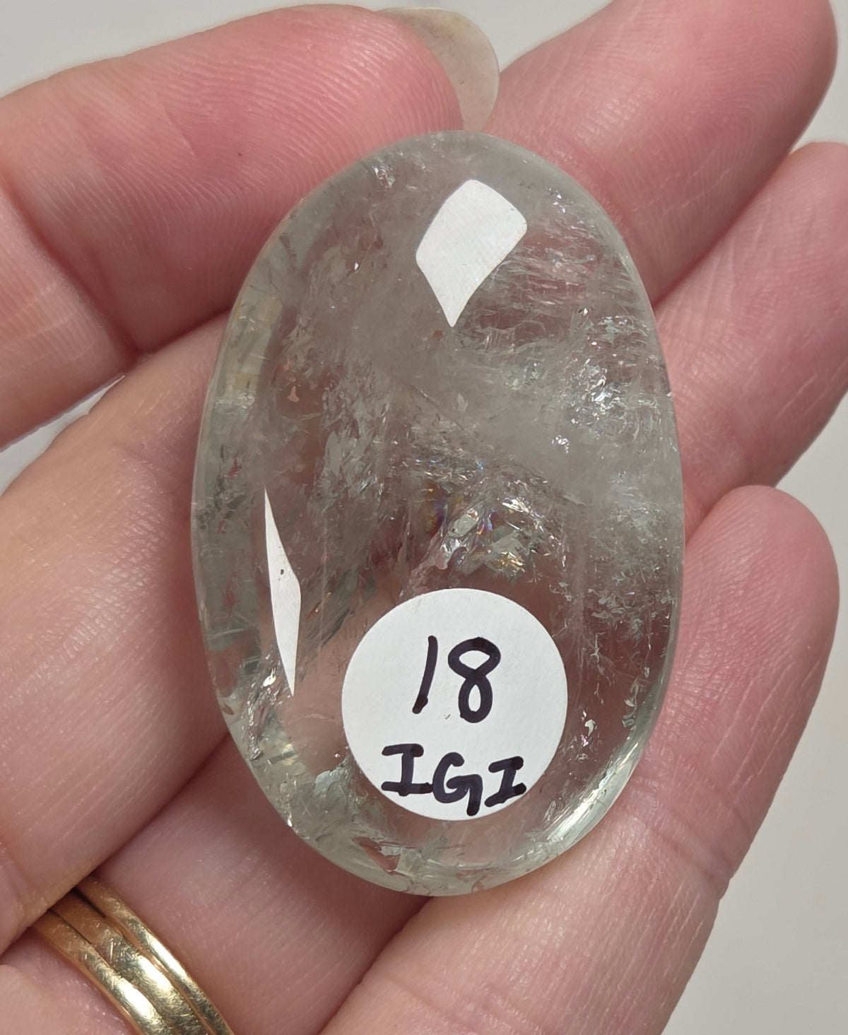 A hand holding a polished, oval-shaped clear gemstone with a white label that reads '18 IGI'.