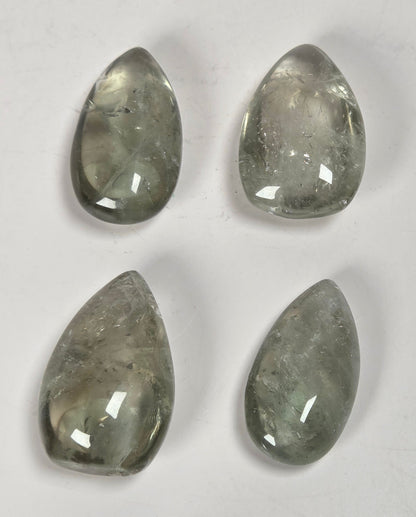 Four teardrop-shaped clear gemstone cabochons with grayish-green internal inclusions, arranged in a 2x2 grid on a light off-white background.