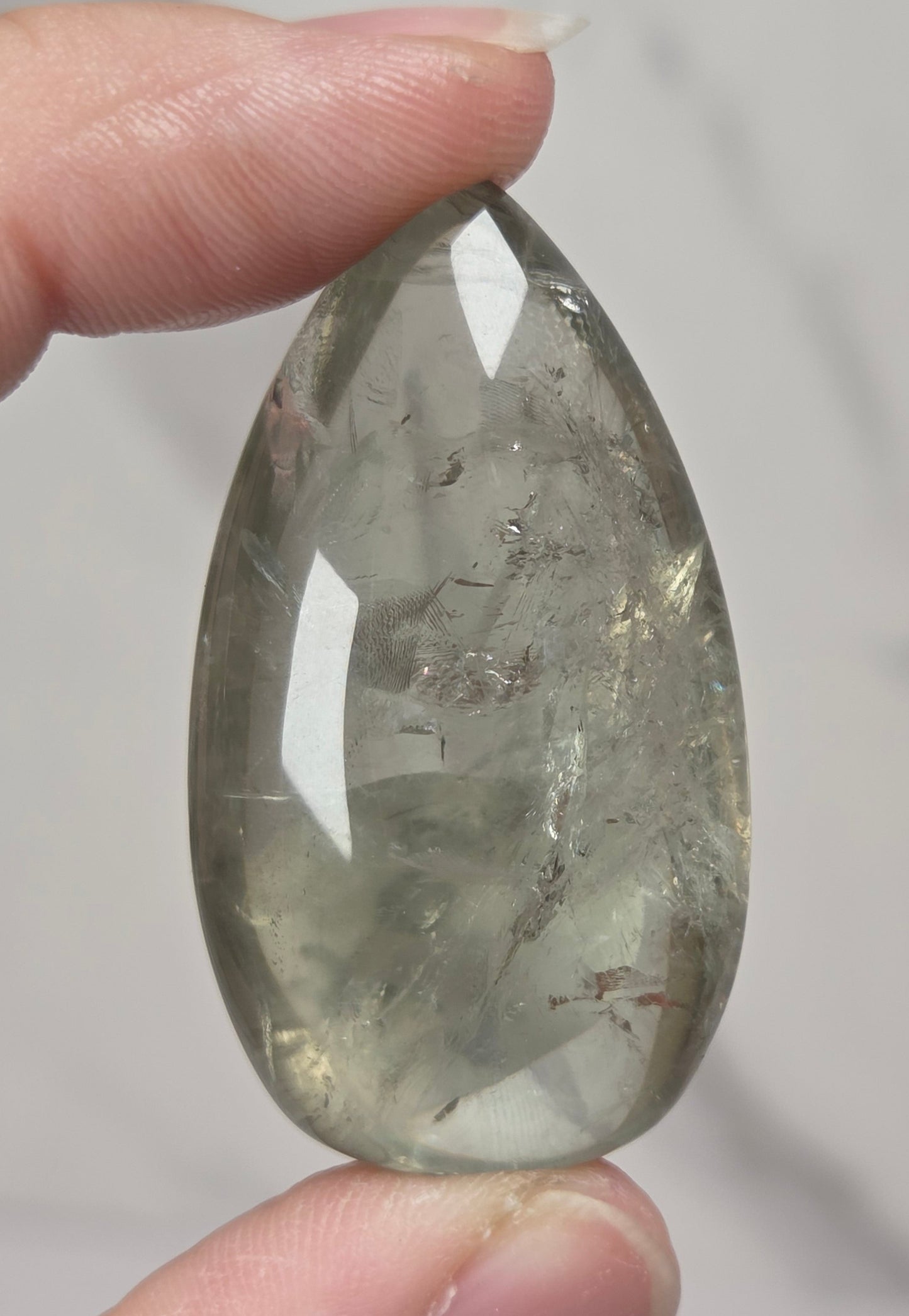 A hand holding a teardrop-shaped clear gemstone with internal inclusions against a light gray marble background.