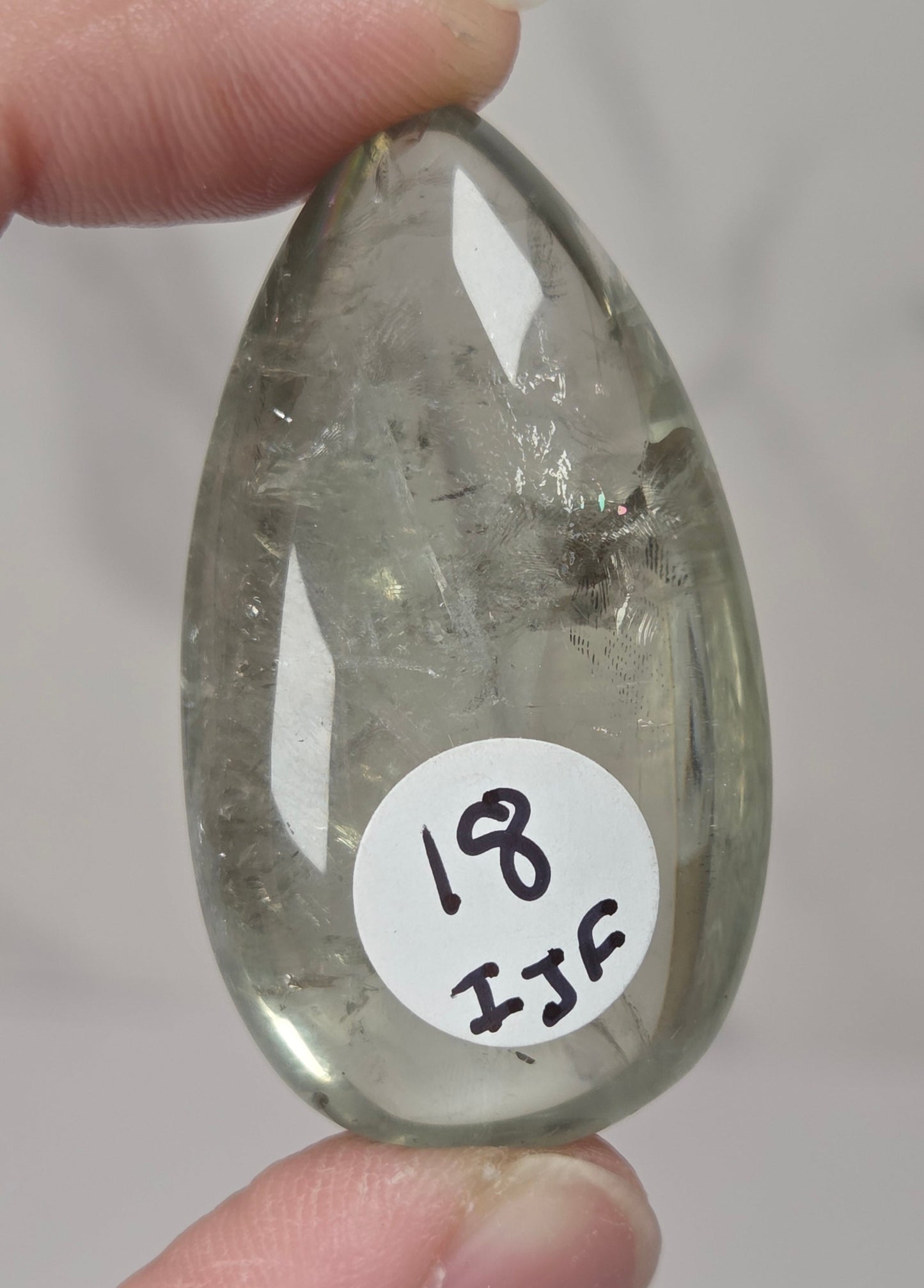A hand holds a polished teardrop-shaped clear stone with visible internal inclusions. A white circular sticker on the stone reads '18 IJF'.