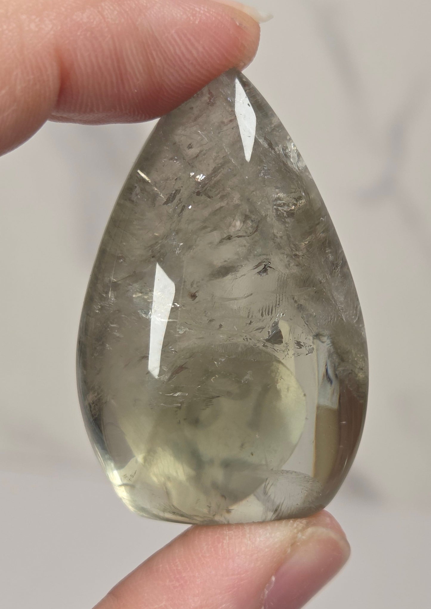 A teardrop-shaped clear gemstone with internal inclusions, held between two fingers against a light gray marble background.
