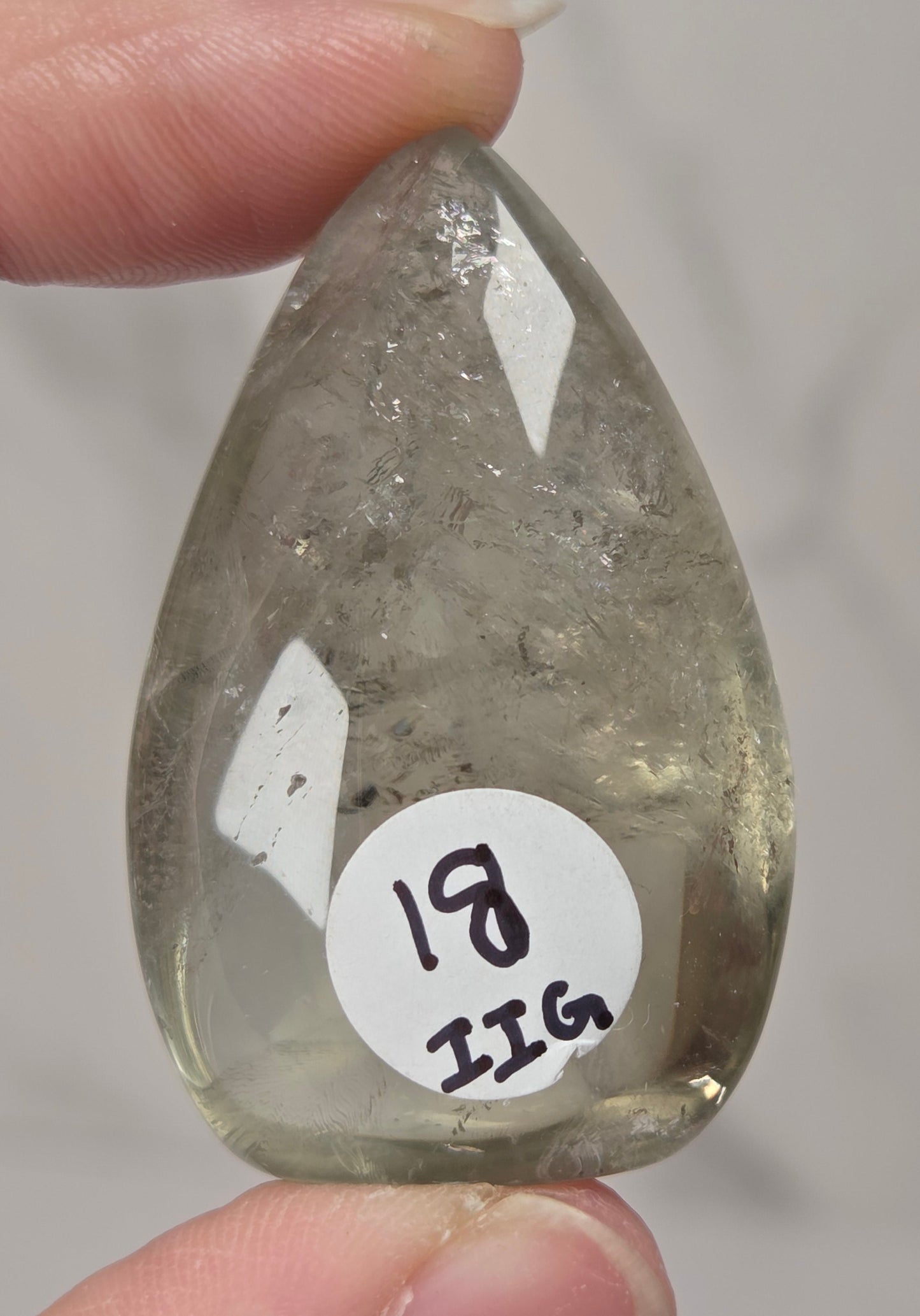 A hand holds a polished teardrop-shaped clear gemstone with internal inclusions and a white circular sticker reading '18 IIG'.