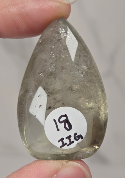A hand holds a polished teardrop-shaped clear gemstone with internal inclusions and a white circular sticker reading '18 IIG'.