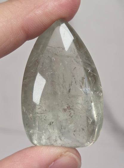 A hand holding a polished, teardrop-shaped clear crystal stone with internal inclusions against a light gray marble background.