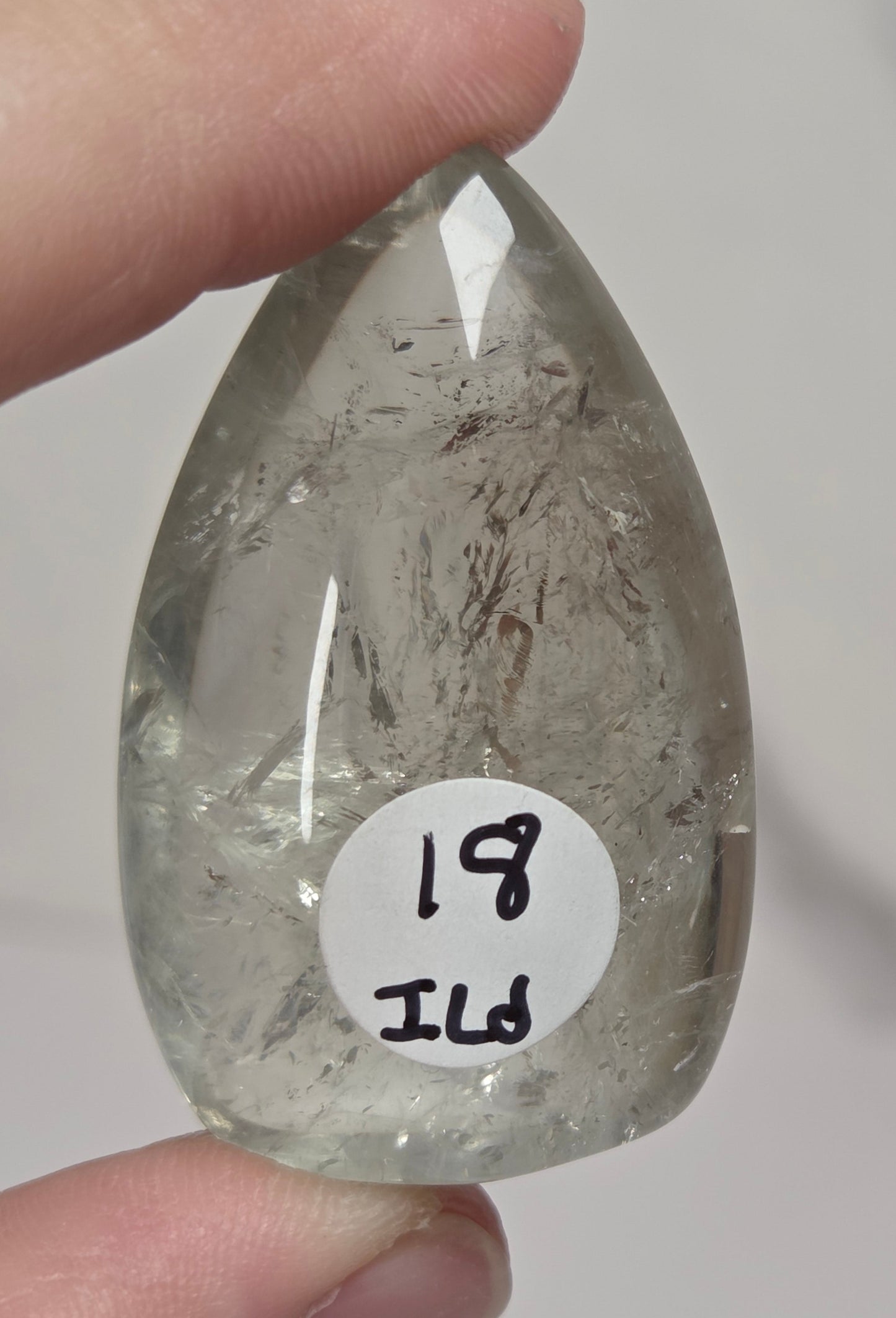 A hand holding a teardrop-shaped clear crystal pendant with internal inclusions and a white circular label reading '19 JUD'.