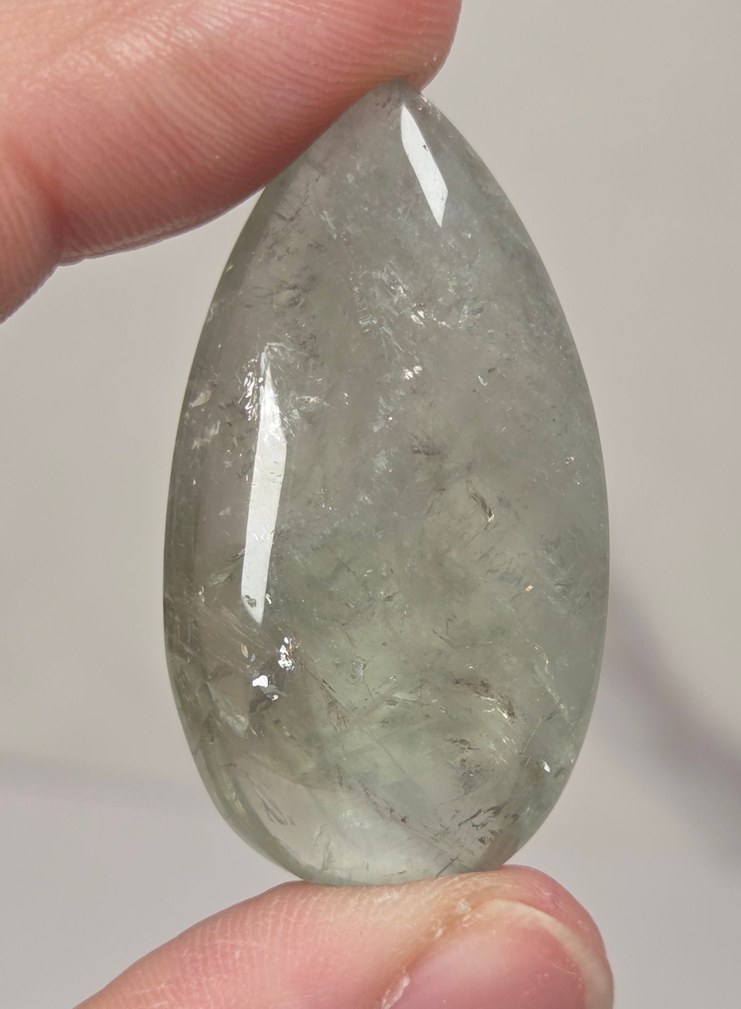 A hand holding a polished teardrop-shaped clear stone with internal inclusions, shown close-up against a light neutral background.