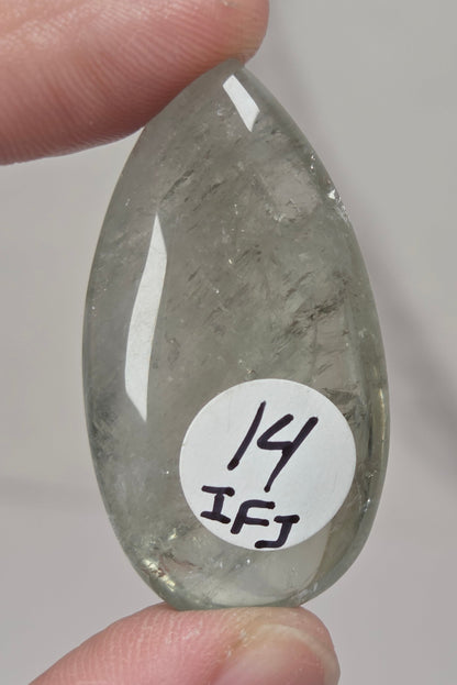 A hand holds a polished, teardrop-shaped gray stone with a white circular sticker that reads '14 IFJ' against a light gray background.