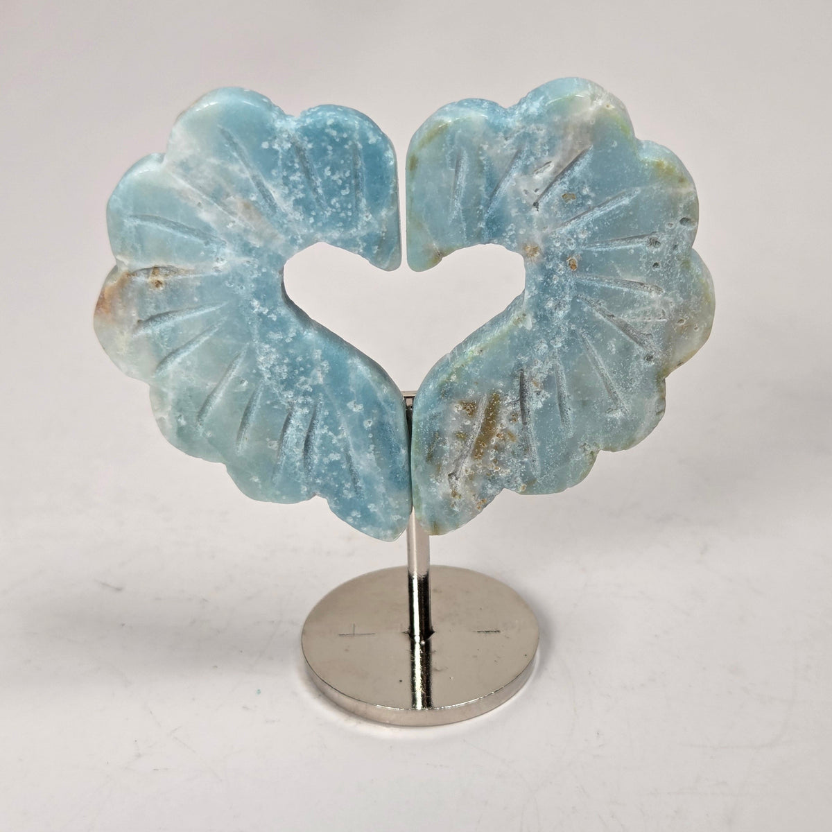 A pair of light blue carved stone earrings shaped like stylized flowers with a heart cutout in the center, displayed on a silver stand against a light gray background.