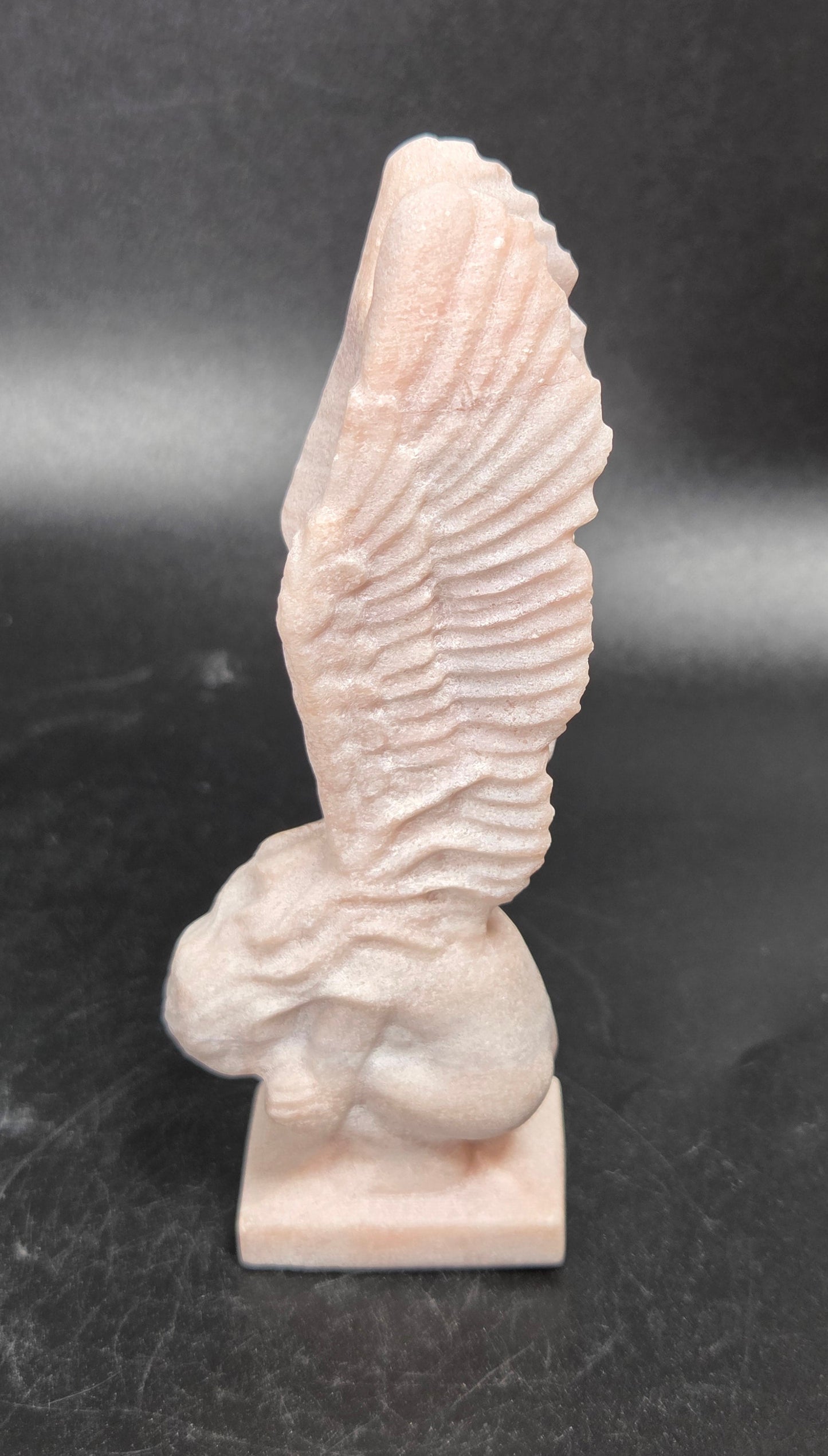 A small pale pink stone sculpture of a winged figure, likely a cherub or angel, standing on a rectangular base against a dark gray background.