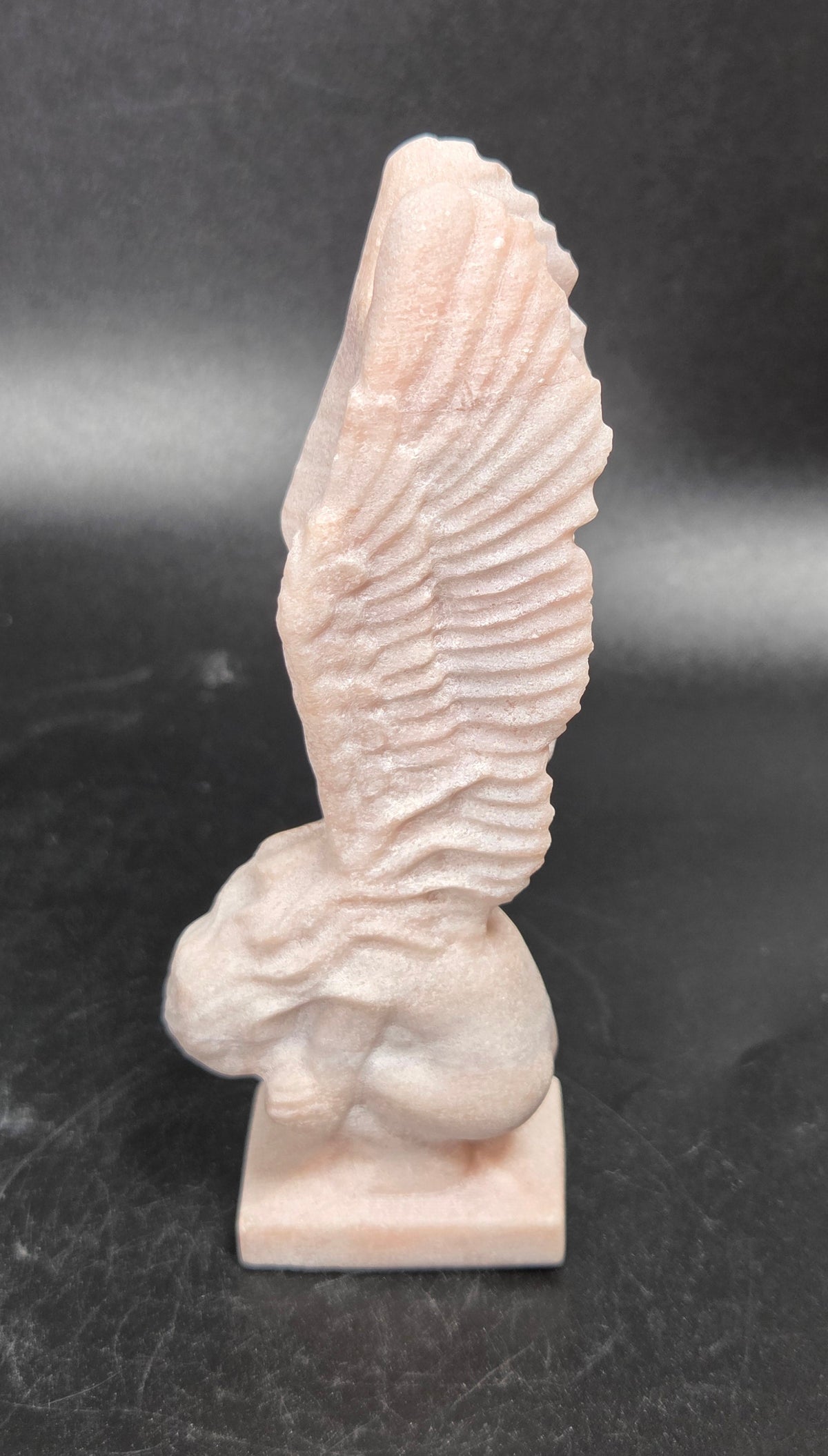 A small pale pink stone sculpture of a winged figure, likely a cherub or angel, standing on a rectangular base against a dark gray background.