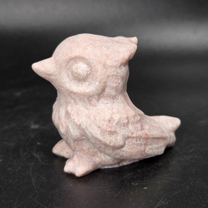A small carved bird figurine, likely made of stone or resin, in a dusty pink color, standing on a dark black surface.