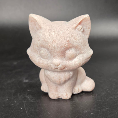 A small pale pink cat figurine with a speckled stone-like texture, sitting on a dark gray surface.