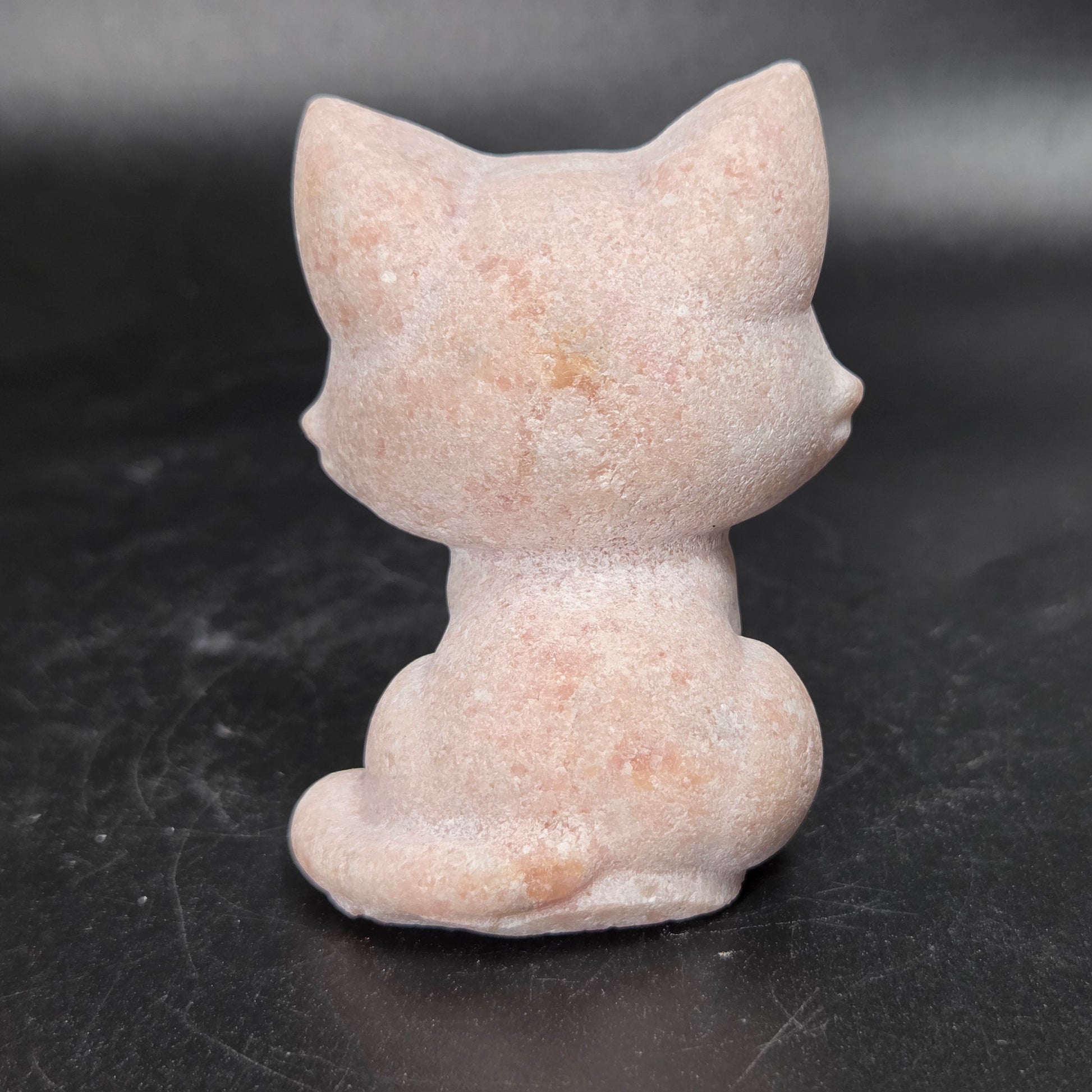 A small pink cat figurine shown from the back, made of a textured stone-like material, set against a dark gray background.