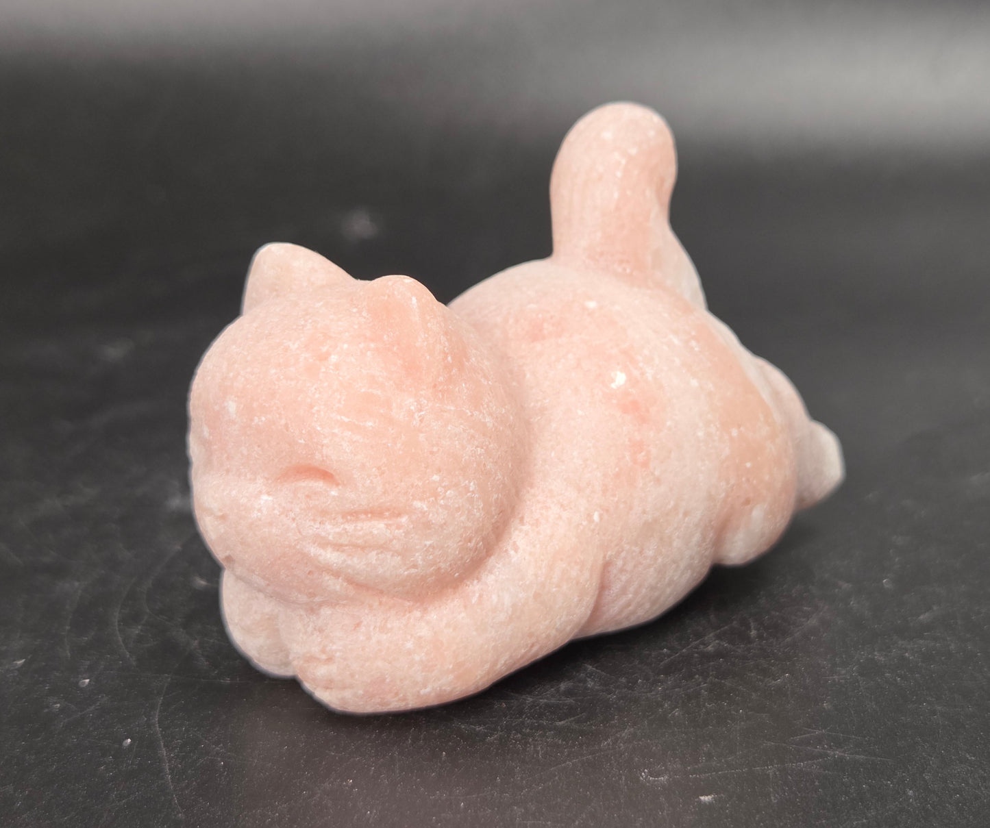 A small pink cat figurine carved from polished stone, lying down on a dark gray surface.