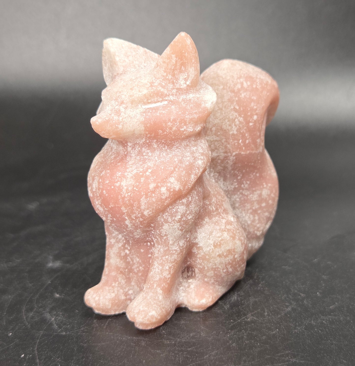 A small carved figurine of a fox made from pale pink stone, standing on a dark gray surface against a dark gray background.