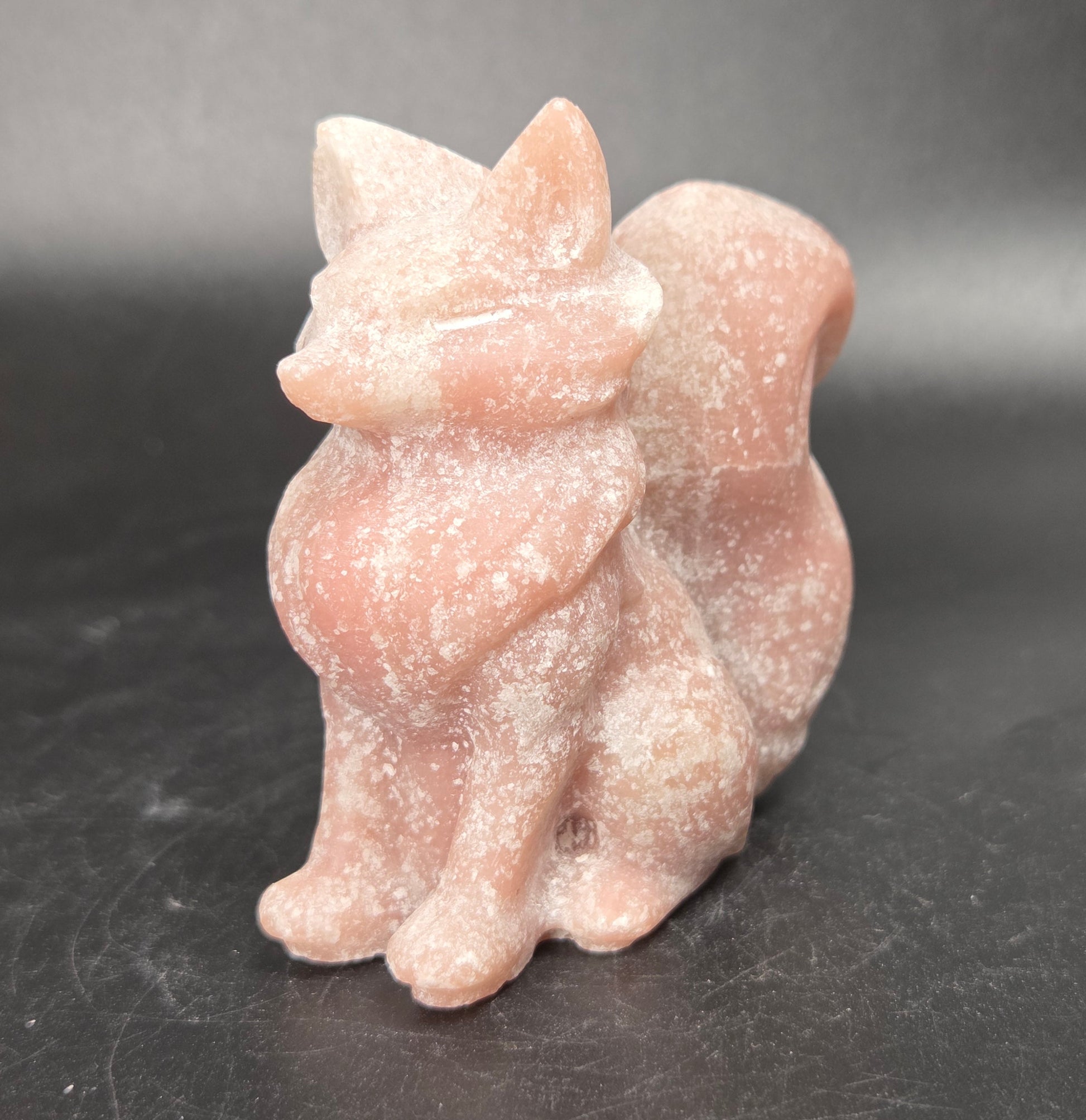 A small carved figurine of a fox made from pale pink stone, standing on a dark gray surface against a dark gray background.