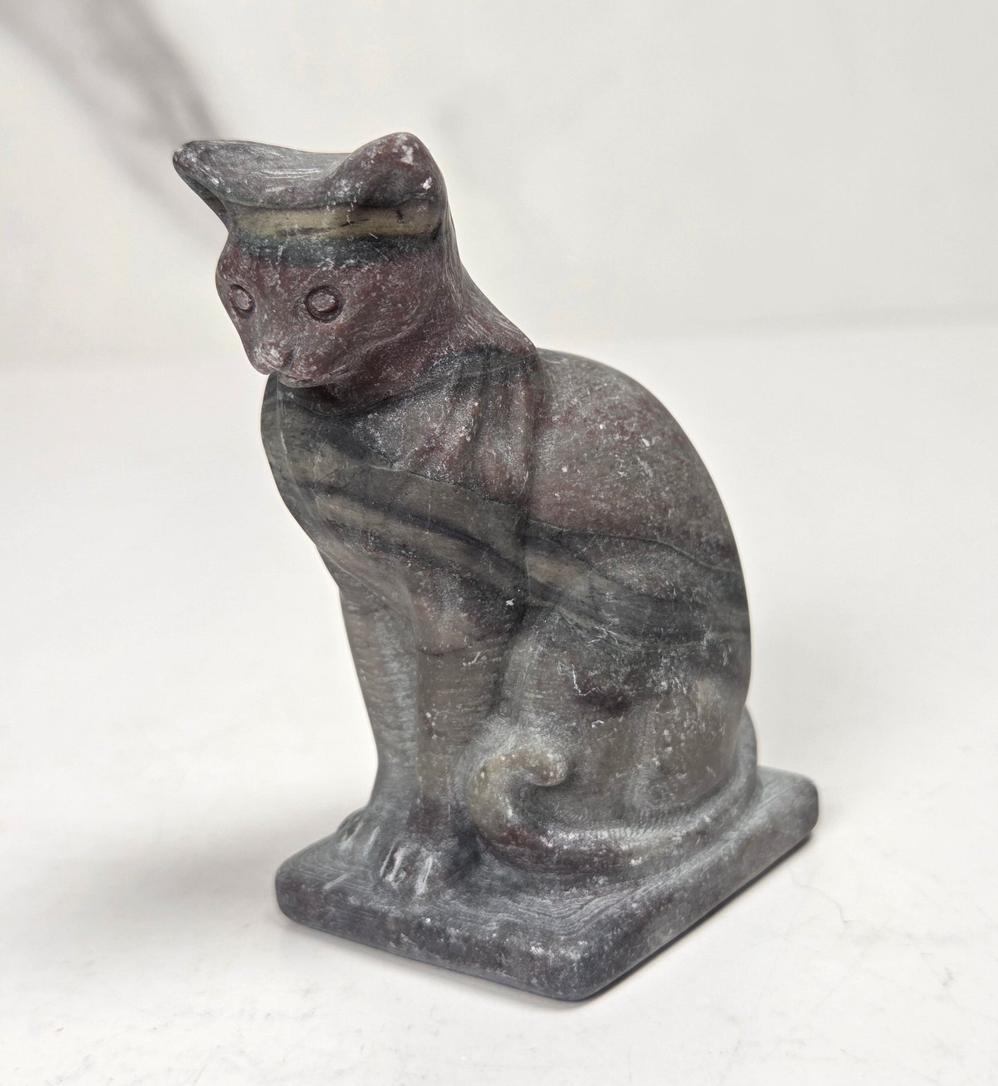 A small carved cat figurine in dark gray stone with subtle brown and greenish patina, standing on a rectangular base against a plain light background.