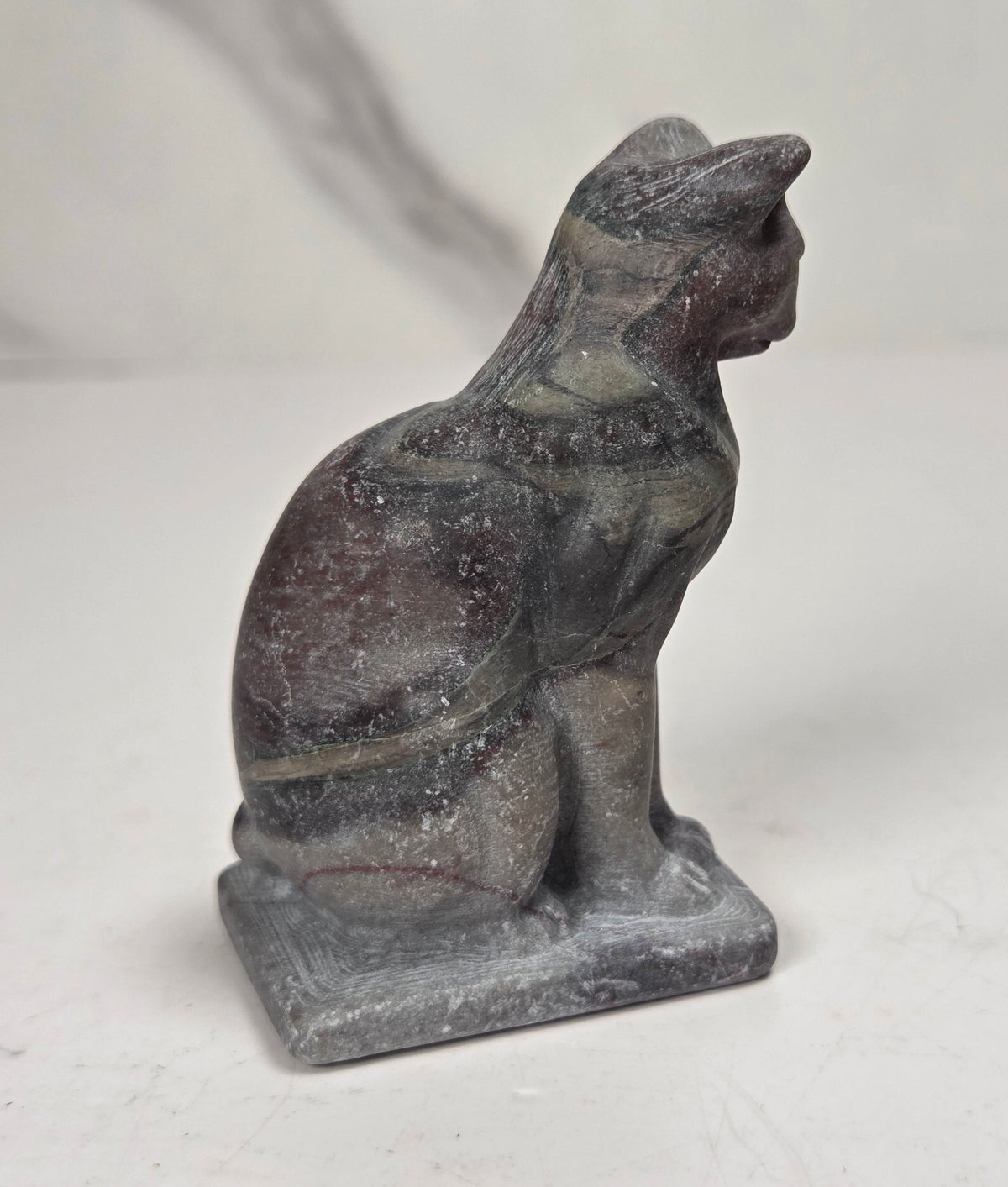 A small carved stone cat figurine, likely made of slate or basalt, shown in profile sitting on a rectangular base against a light gray and white marble-patterned background.