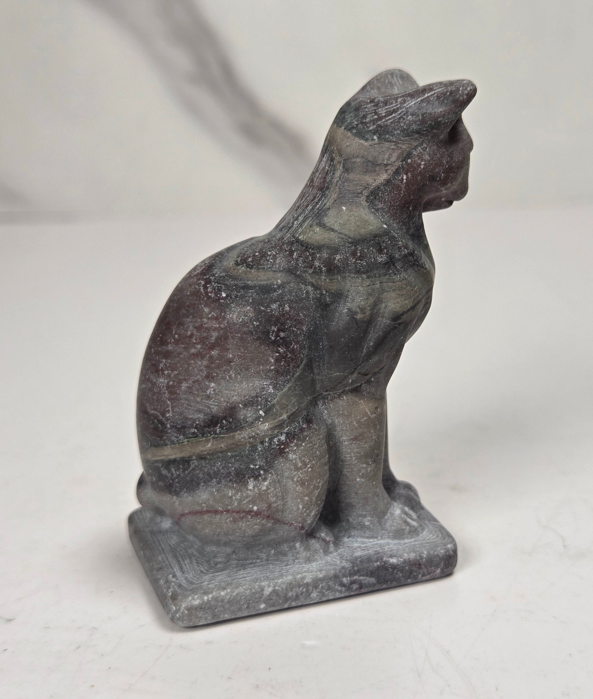 A small carved stone cat figurine, likely made of slate or basalt, shown in profile sitting on a rectangular base against a light gray and white marble-patterned background.