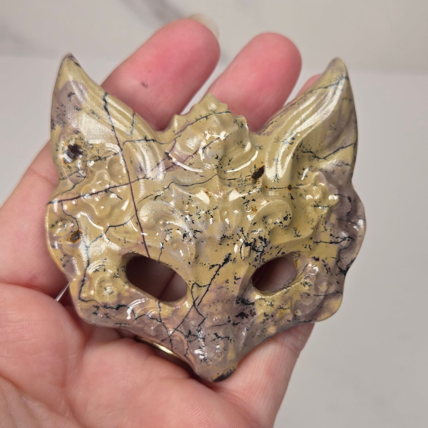 A hand holding a small carved mask or ornament with a textured, organic surface in beige, brown, and gray tones, featuring two eye openings and pointed ear-like protrusions.